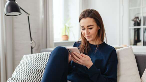 Frau (mittleren Alters) sitzt auf dem Sofa und liest mir einem kleinem Lächeln etwas auf ihrem Handy