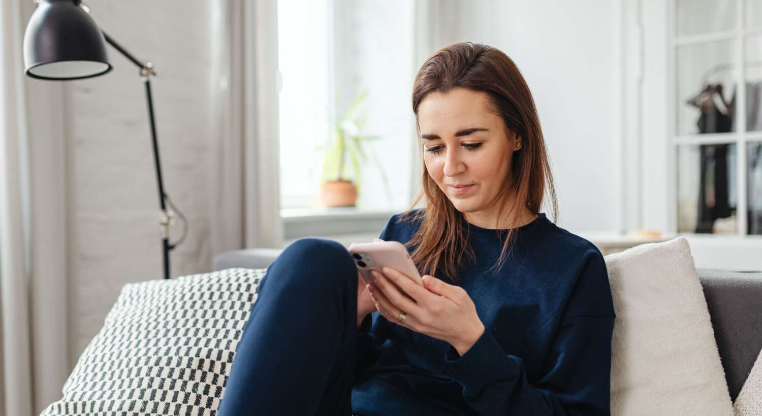 Frau (mittleren Alters) sitzt auf dem Sofa und liest mir einem kleinem Lächeln etwas auf ihrem Handy